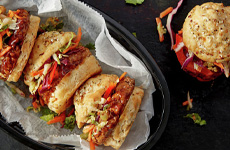 Basket with white deli paper filled with three Korean BBQ turkey burgers, BBQ sauce, and coleslaw on biscuit buns with one biscuit burger on a plate on the side.