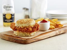 Spicy Chicken Biscuit Sandwich recipe on wooden cutting board platter with fries, ketchup, a beer bottle, and condiments in background.