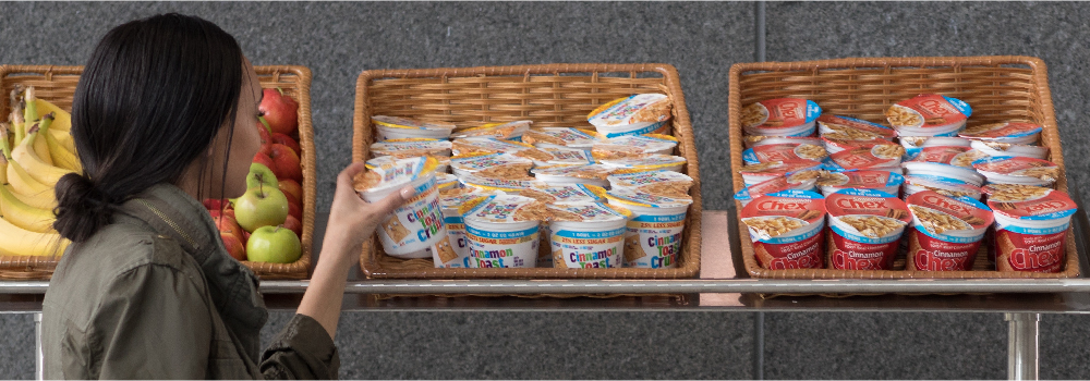 K-12 student selects cereal from a basket in school cafeteria. Fresh whole fruit is also shown.