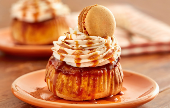 A plated pumpkin spice cinnamon roll topped with vanilla cream and dripping with caramel sauce, topped with a macaron.