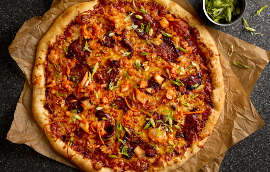 A freshly baked pizza topped with kimchi, sliced green onions, and a variety of other colorful toppings, placed on crinkled parchment paper over a dark