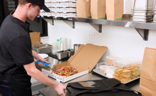 Person with black hat cutting pizza inside pizza box at prep station next to delivery bag.